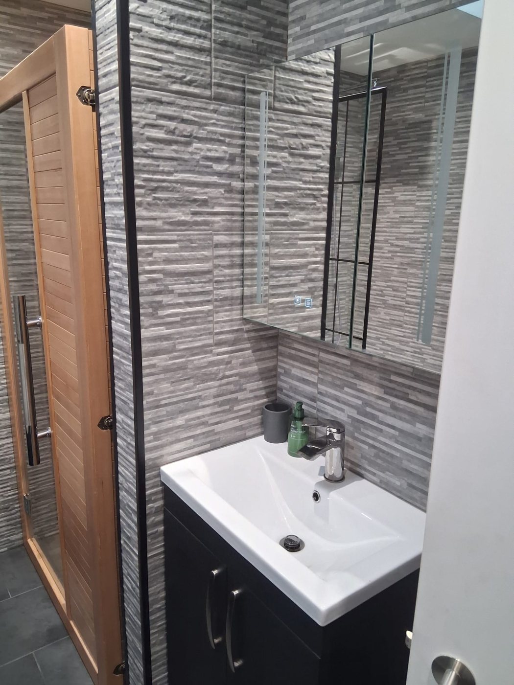 White basin on a dark vanity unit below a mirrored cabinet, with the sauna door alongside