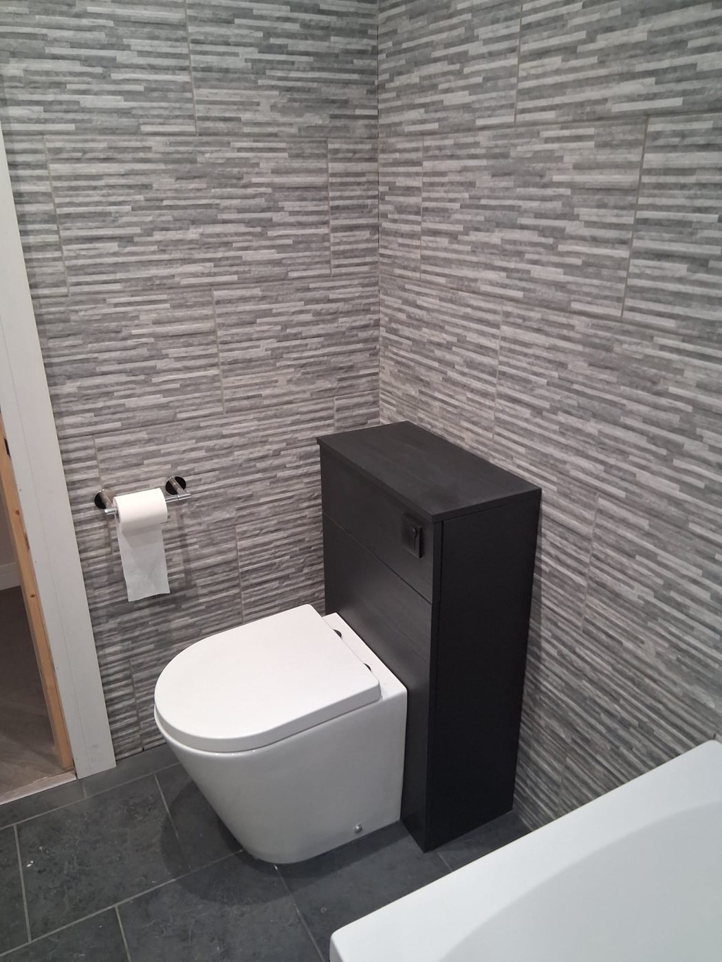 Back-to-wall toilet with a dark cistern unit against grey split-face effect wall tiles and a slate grey tiled floor