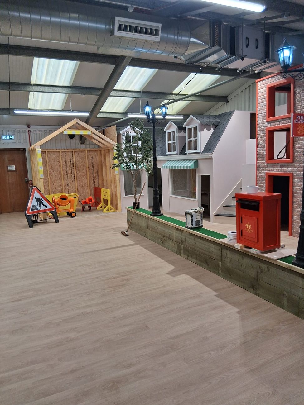 Indoor play village street with pitched-roof shop fronts, a green awning and a road-works corner with cones and a mixer